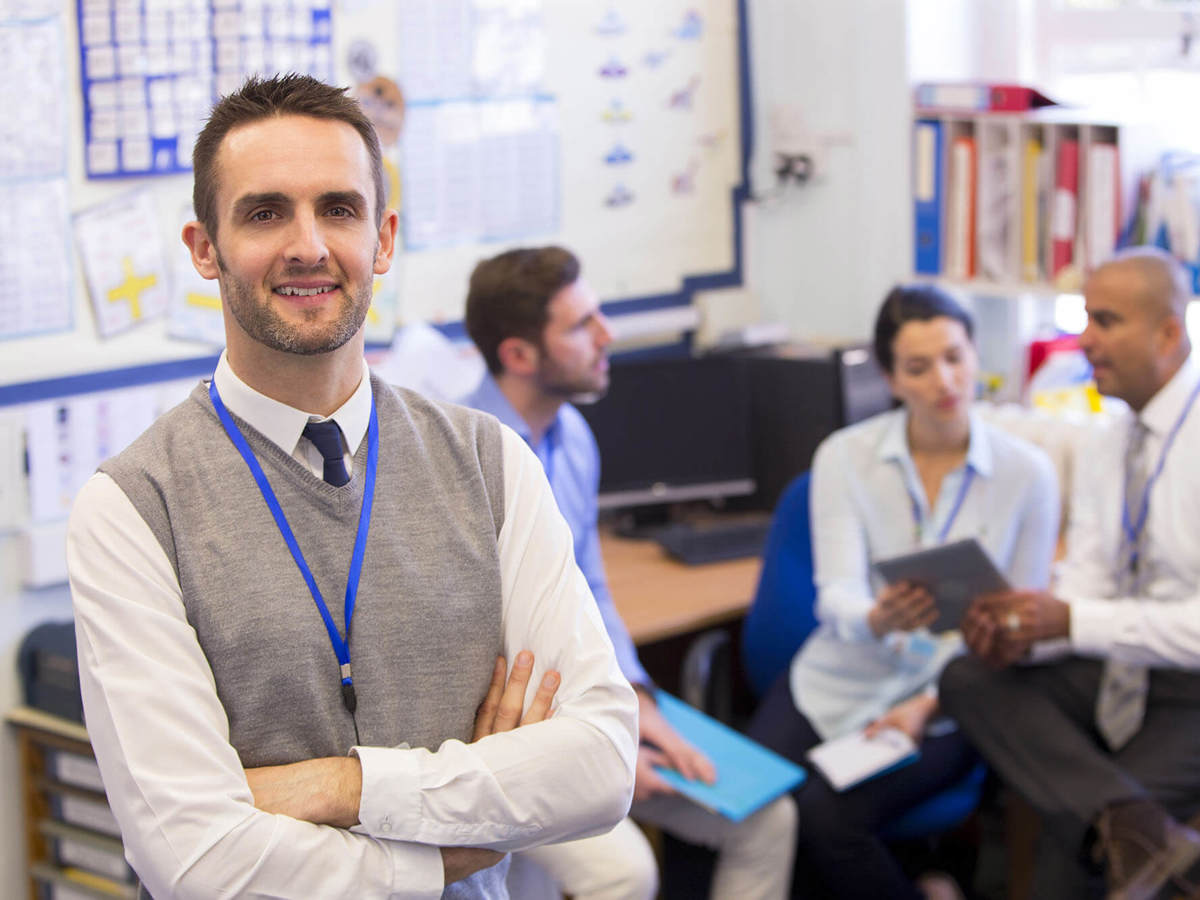 people teacher smiling into the camera with other teachers talking to each other behind him